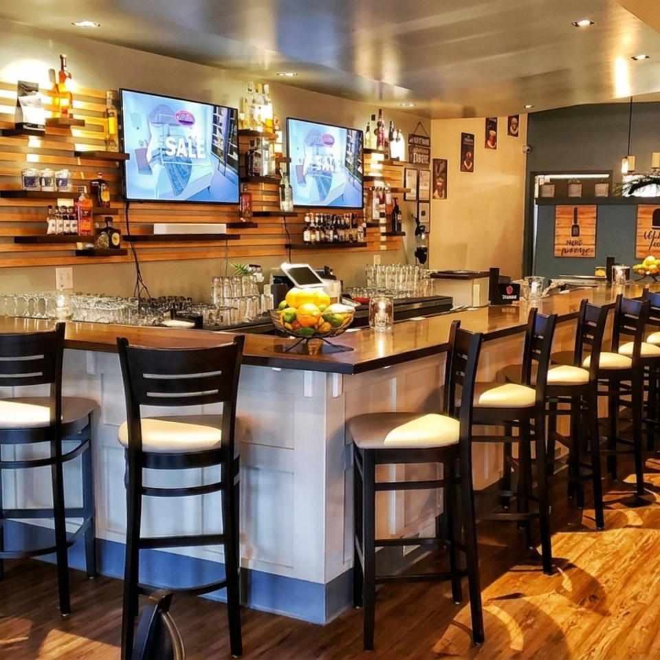 Warmly lit restaurant bar with high chairs, wooden floors, cocktail bottles, glassware, fruit centerpiece, and wall-mounted televisions.